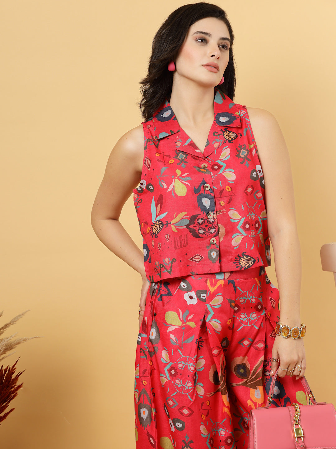Woman in a red floral top & matching wide Leg pants outfit holding a pink handbag against a beige background.
