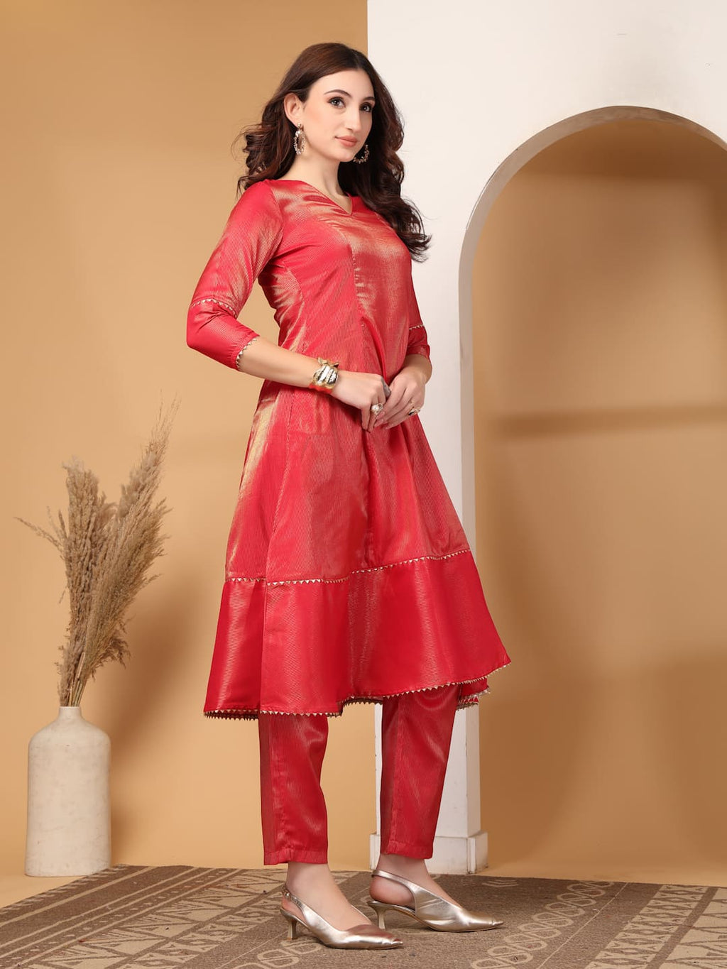 Woman wearing a festive red flared A-line kurti with vertical gold metallic stripes, delicate gota lace trimming, and matching red trousers. Side View