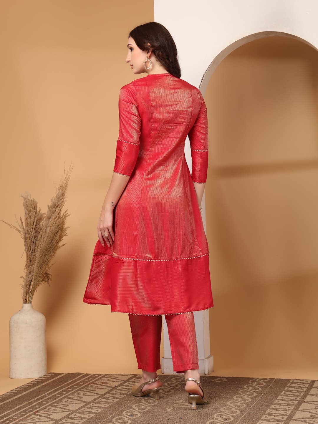 Woman wearing a festive red flared A-line kurti with vertical gold metallic stripes, delicate gota lace trimming, and matching red trousers. Back View