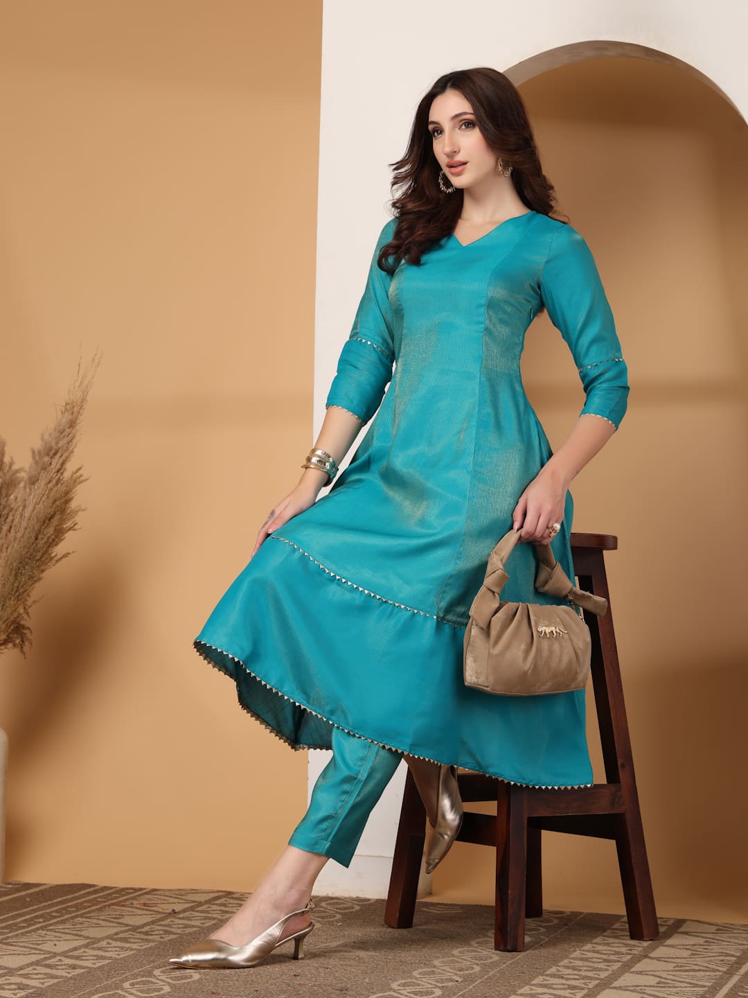 Woman wearing a festive turquoise colour flared A-line kurti with vertical gold metallic stripes, delicate gota lace trimming, and matching turquoise trousers. Style Pose.