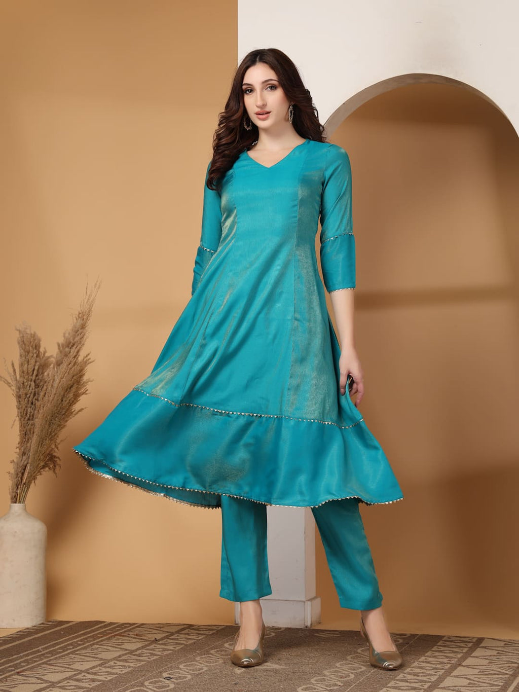 Woman wearing a festive turquoise colour flared A-line kurti with vertical gold metallic stripes, delicate gota lace trimming, and matching turquoise trousers. Front View