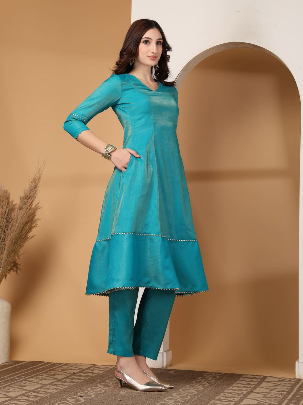 Woman wearing a festive turquoise colour flared A-line kurti with vertical gold metallic stripes, delicate gota lace trimming, and matching turquoise trousers. Side View