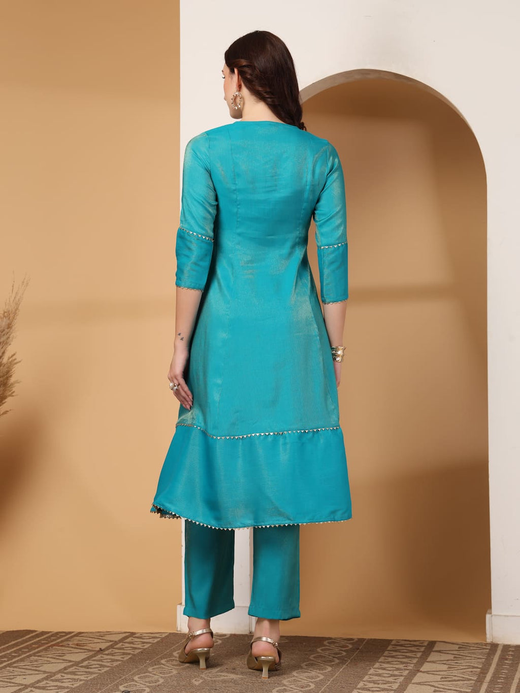 Woman wearing a festive turquoise colour flared A-line kurti with vertical gold metallic stripes, delicate gota lace trimming, and matching turquoise trousers. Back View