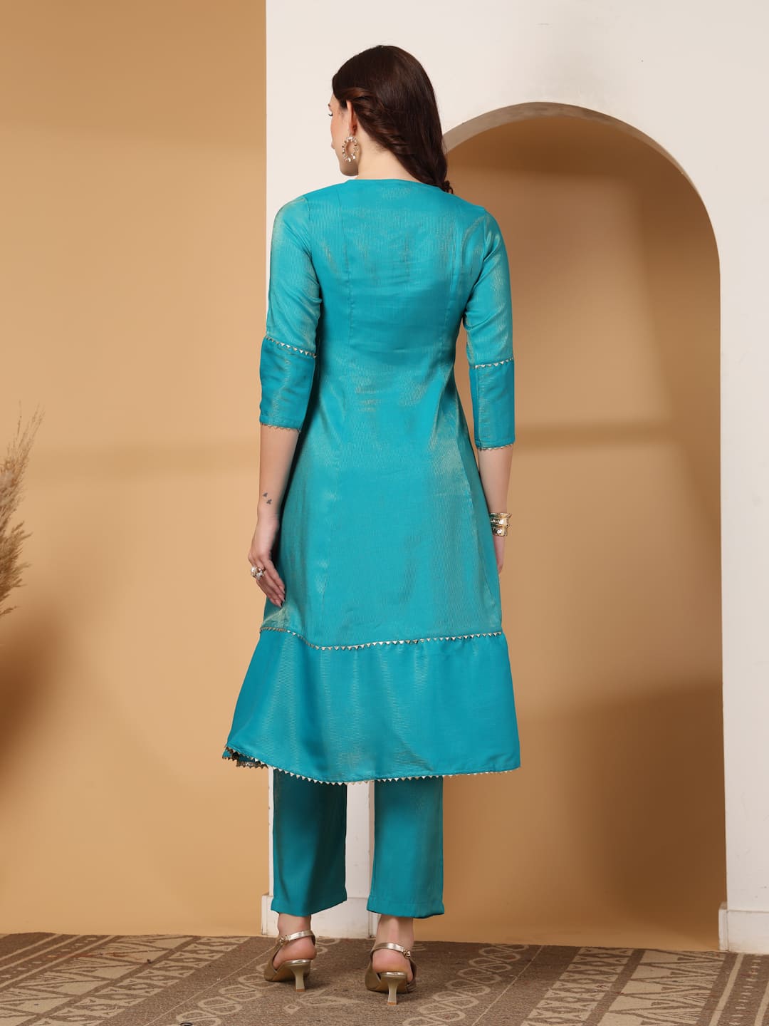 Woman wearing a festive turquoise colour flared A-line kurti with vertical gold metallic stripes, delicate gota lace trimming, and matching turquoise trousers. Back View